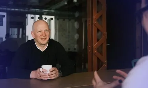 Man in black sweater holding a white mug, engaged in conversation with another person whose hands are visible.