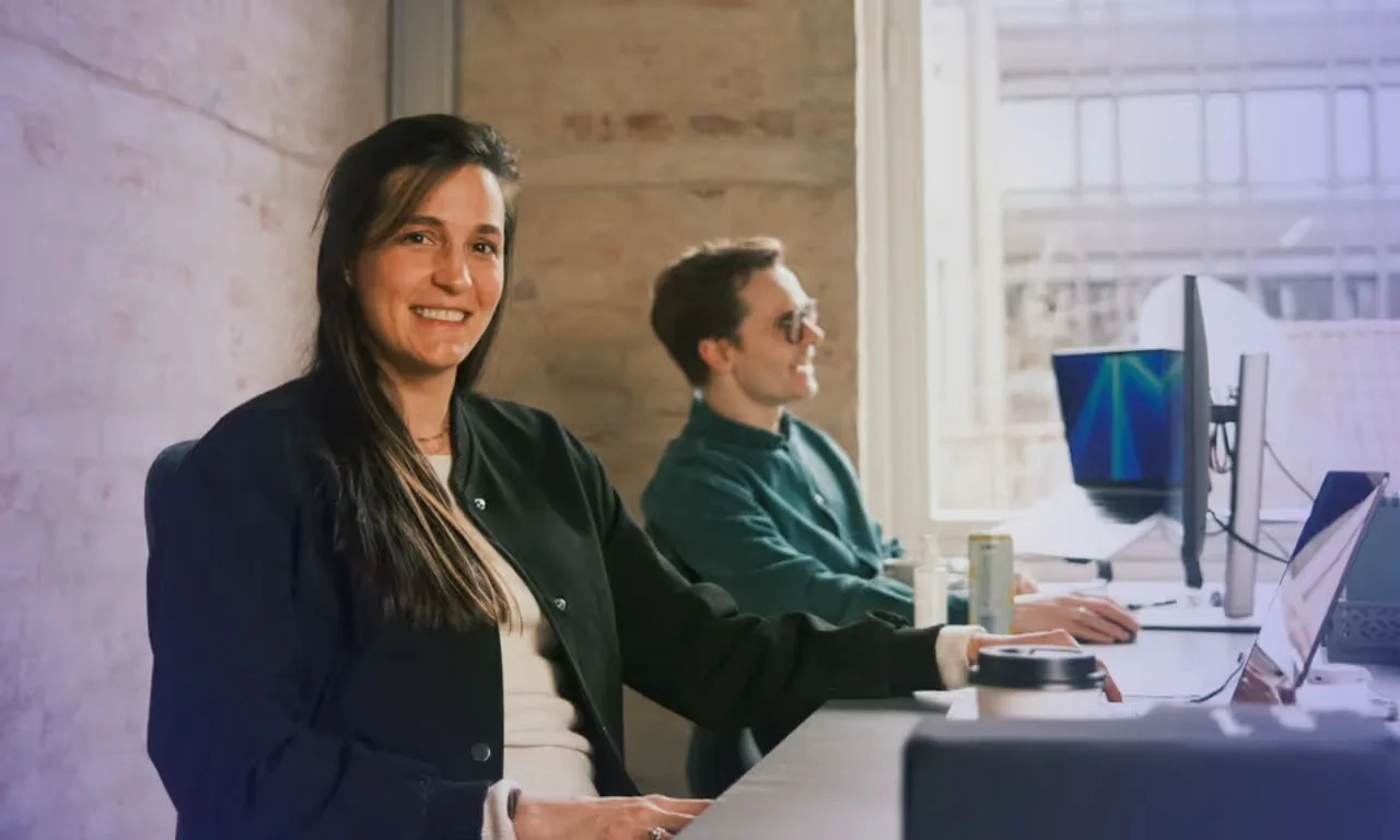 Two colleagues working at desks facing computers in a bright office; a woman in the foreground smiles at the camera.
