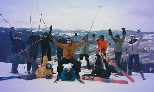 Group of twelve skiers posing cheerfully on a snowy mountain slope with ski poles raised, scenic mountainous landscape in the background.