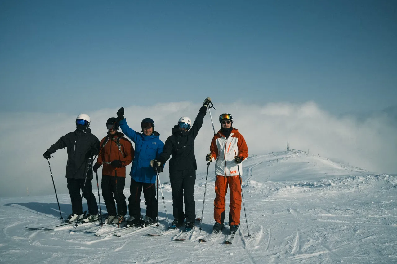 Group of five skiers standing on snow-covered slope with ski gear under a clear blue sky.